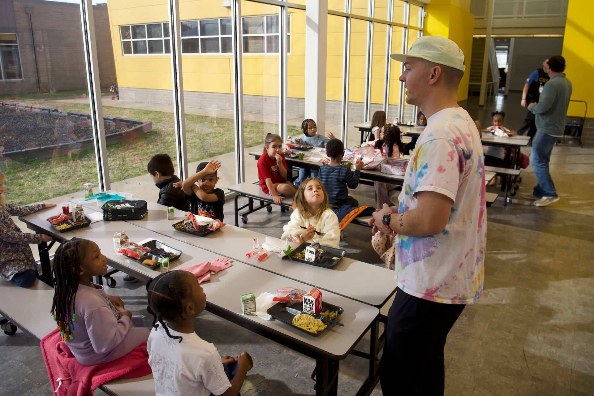 Copy of P1111605 (1) A man stands and talks to a group of young children sitting at cafeteria tables eating lunch in a bright room with large windows and yellow walls.