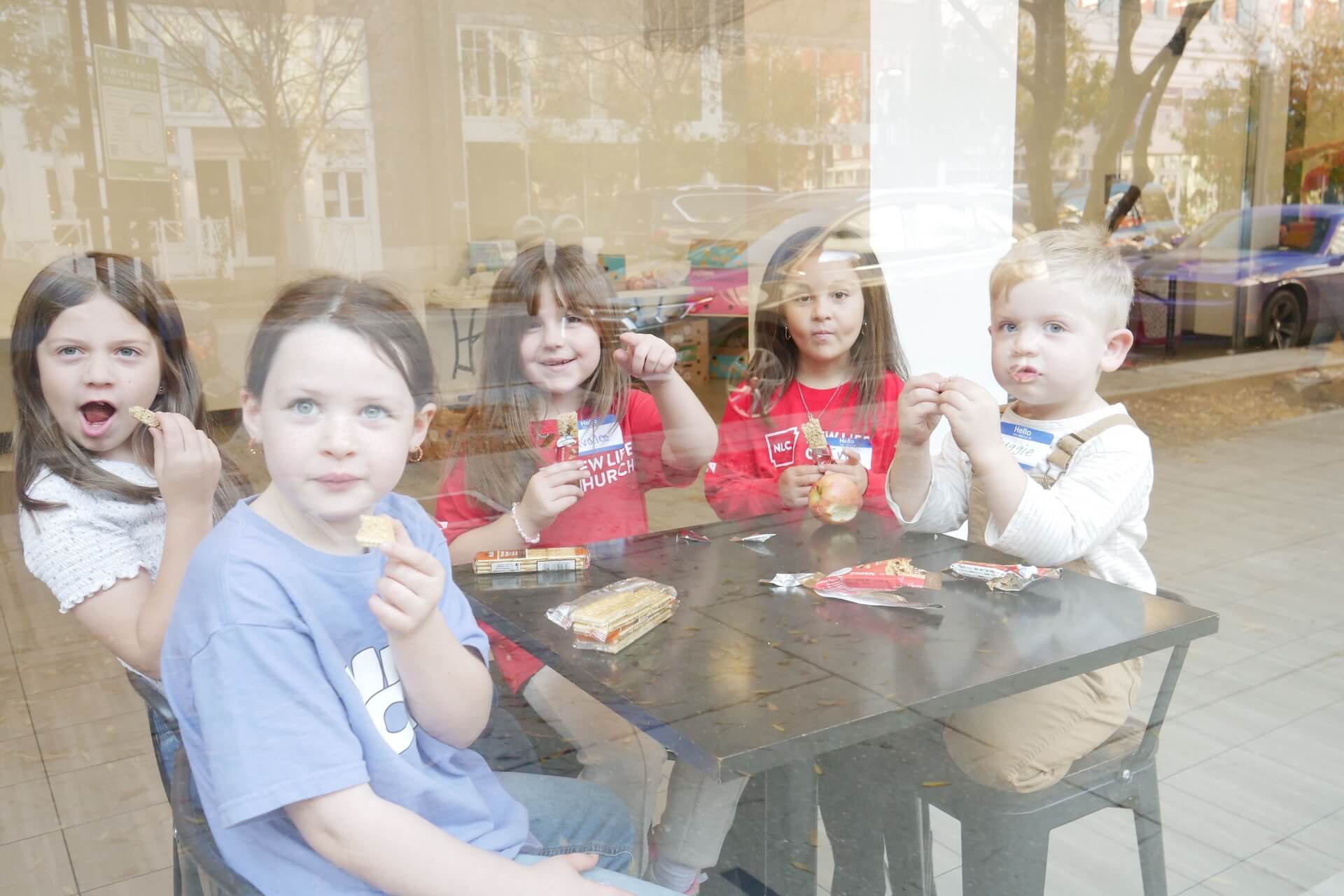 Five young children sit around a black table by a window, eating snacks and looking toward the camera. Packaged snacks and drinks are on the table; reflections from the glass partially obscure the scene.