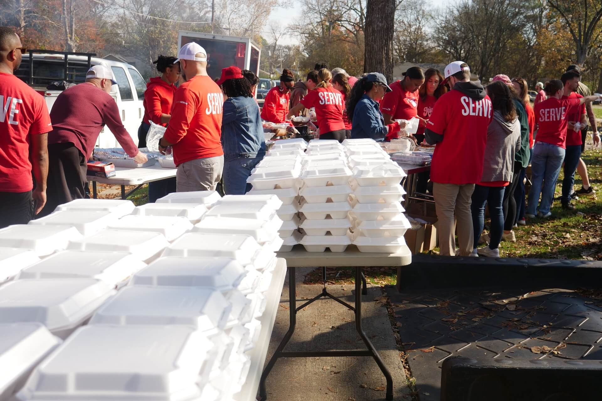 A group of people in red shirts labeled SERVE prepares and distributes food in white containers outdoors, with tables and stacks of containers visible.