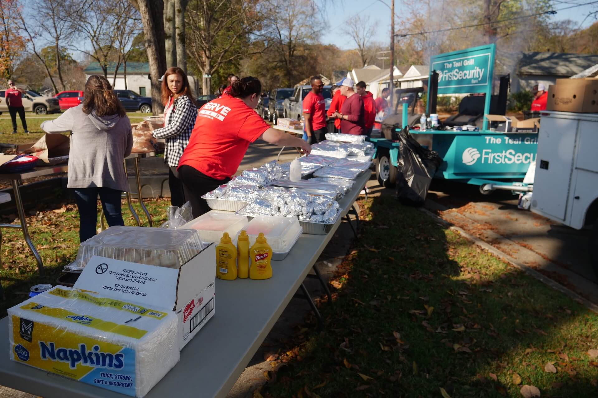 Volunteers in red shirts prepare food at an outdoor catering table near a FirstSecurity branded food trailer.