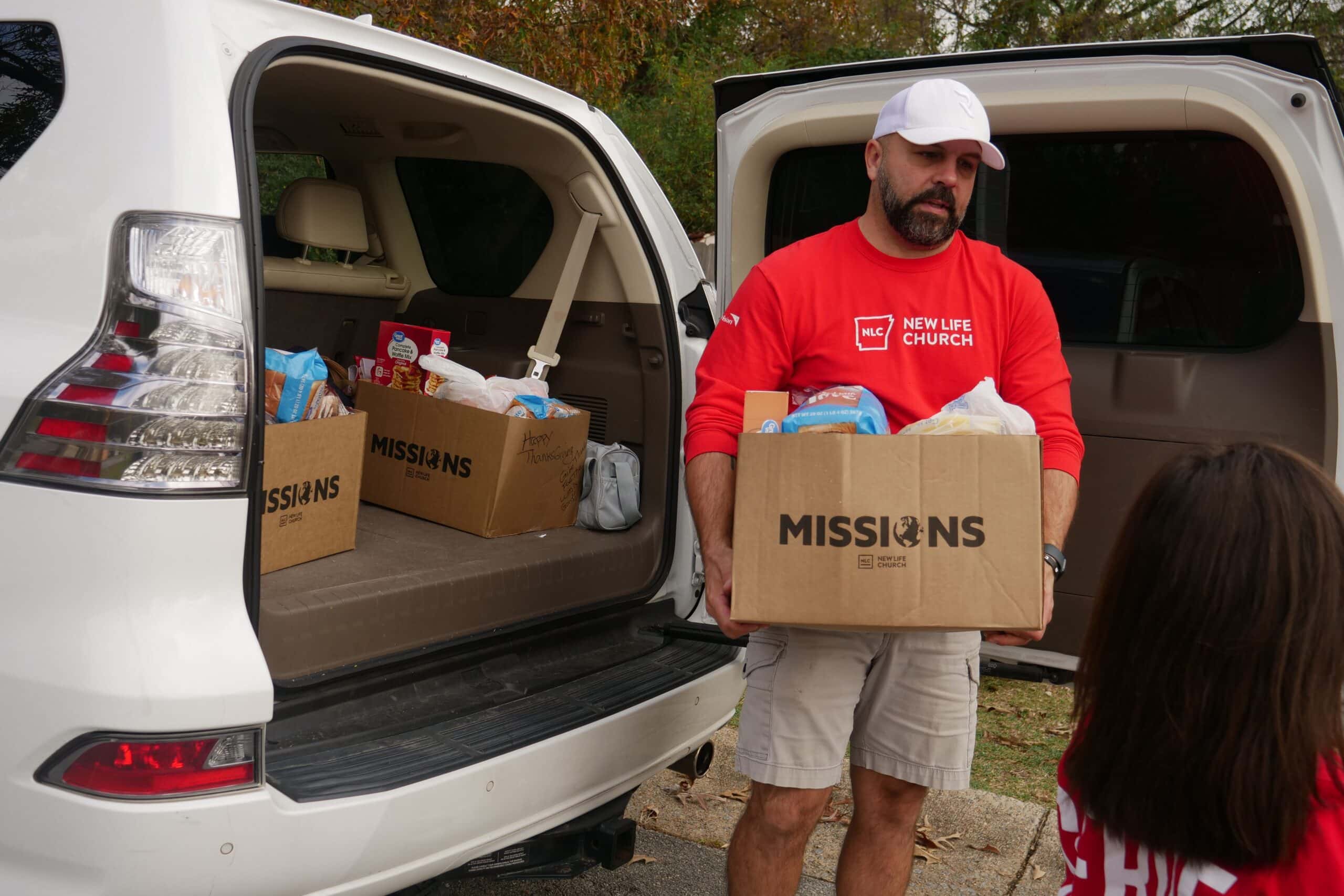 A man wearing a New Life Church shirt hands a cardboard box labeled Missions to a person near the open trunk of a white SUV filled with boxes of food.